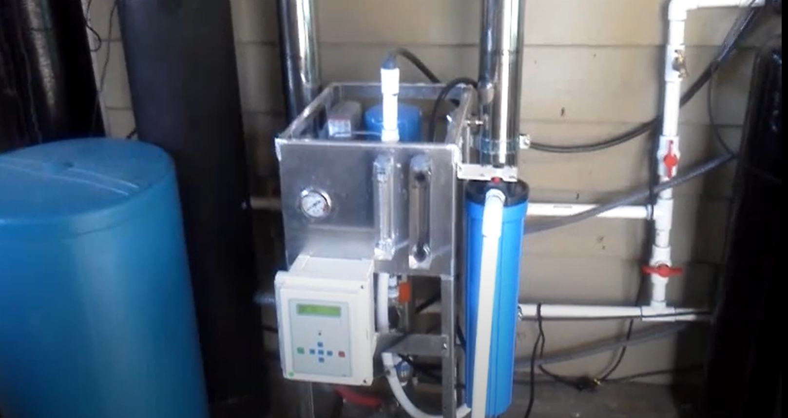 Image of an industrial whole house water filtration system, featuring a network of pipes, a blue hose, and an electronic control panel with various buttons and indicator lights. A large blue barrel is positioned to the left, and the system is housed within a room with concrete walls and additional piping visible in the background, indicative of a water treatment facility.